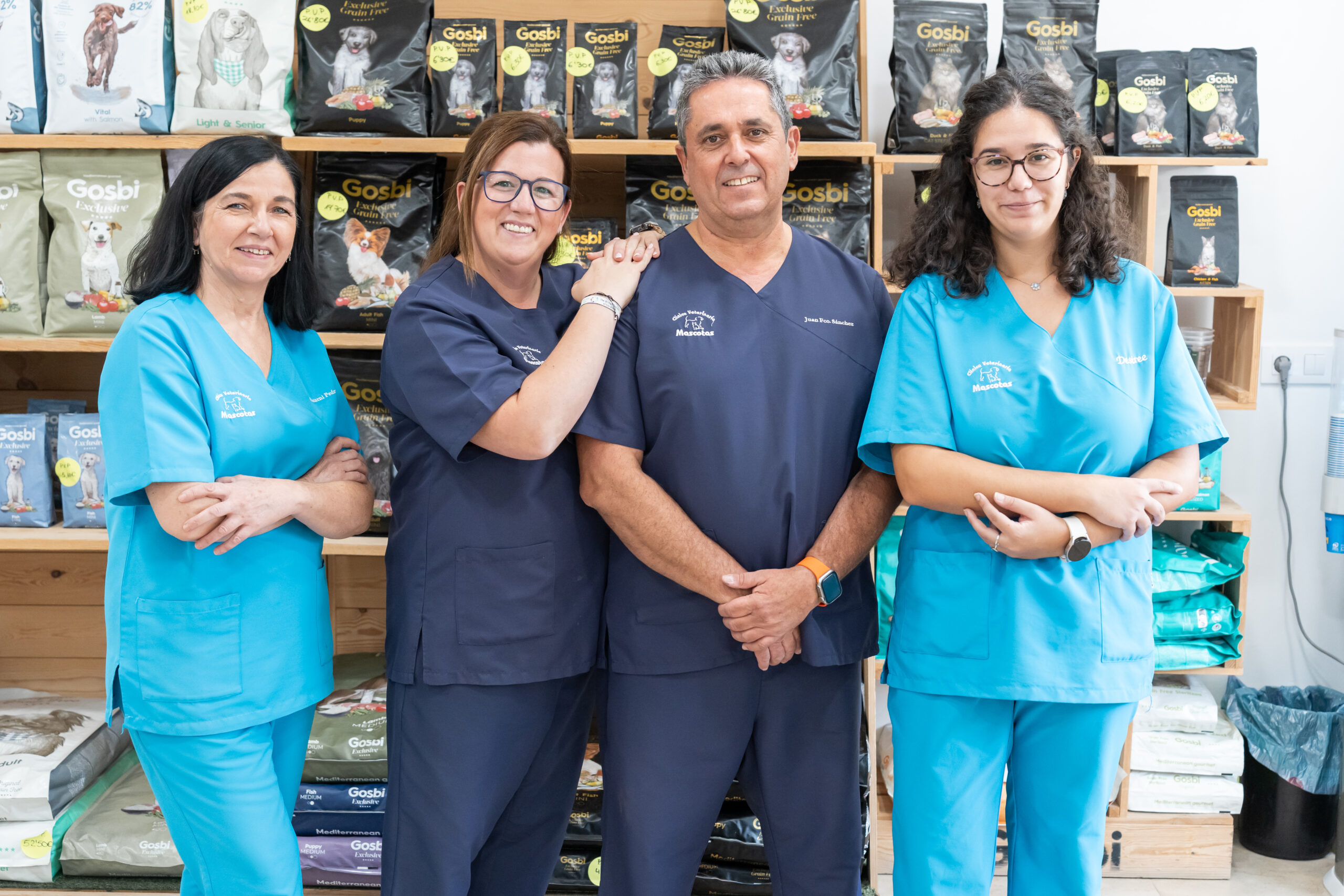 _DSC6664 Four smiling veterinary professionals in scrubs stand together at the Clínica Veterinaria, in front of shelves stocked with pet food and supplies. They pose confidently—two women in blue scrubs, two in navy—arms crossed or hands on shoulders.